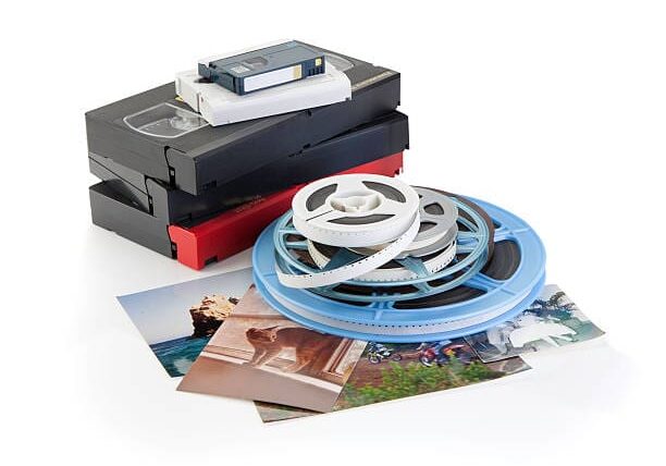 A stack of VHS tapes and a compact cassette next to film reels and scattered photographs on a white background.