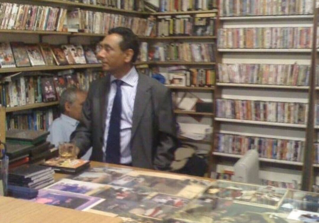 A man in a suit stands behind a counter in a shop filled with shelves of VHS tapes. Another person sits behind him, partially obscured. The counter is covered with photos and various items.